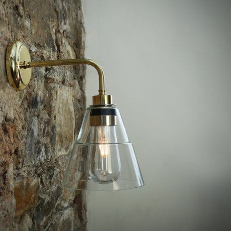 Isvana Glass Cone Wall Light IP65 in polished brass with a clear glass cone shade on a right-angle arm and round wall plate, mounted on a stone wall.