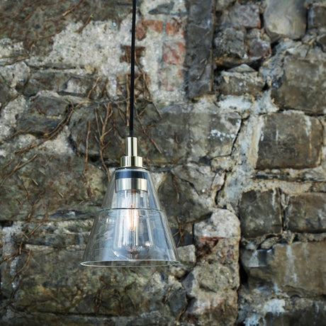 Isvana glass pendant light with stepped cone clear glass shade and polished brass fitting, black cable, shown against a rustic stone wall.