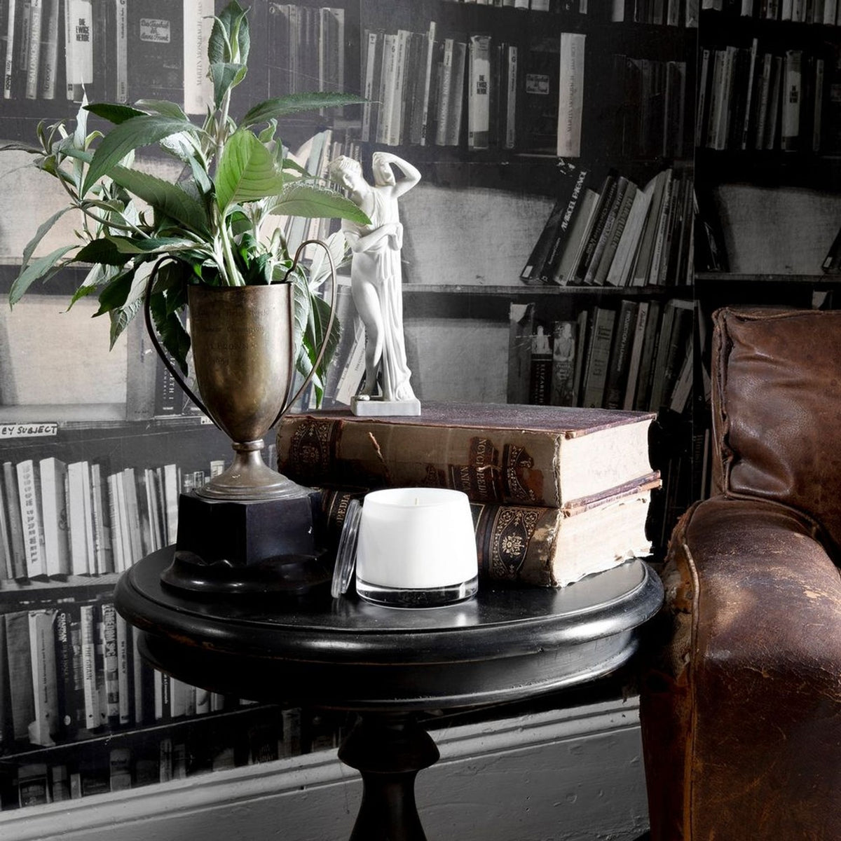 Uplift Sweet Lime & Mandarin scented candle in a white jar with clear lid, displayed on a dark side table beside books, a trophy vase, and a statuette.