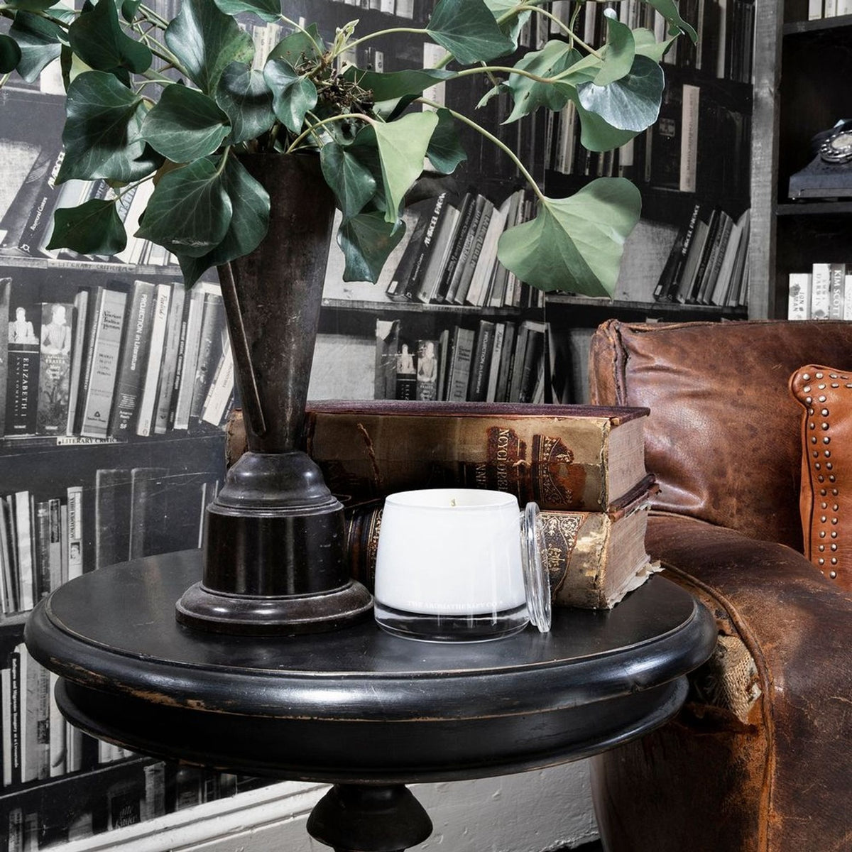 Uplift Sweet Lime & Mandarin Scented Candle 260g in a white jar with clear lid, displayed on a black side table beside vintage books and a leafy plant.
