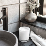 Neroli & Amber Wood Scented Candle in a round white 400g vessel, lid removed, displayed on a dark bathroom shelf beside a stoneware vase and white cloth.