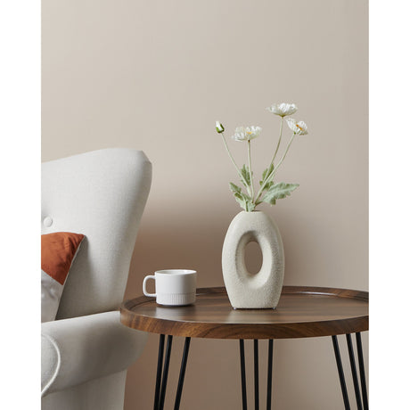 Iwate Ceramic Vase 21cm in beige, a flat oval shape with an open centre, holding dried white flowers on a wooden side table beside a white armchair.