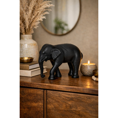 Jabonga Elephant Ornament in black aluminium, shown mid-stride with trunk curled low, on a wooden sideboard beside books, a candle and a vase of dried grasses.