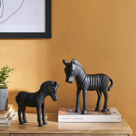 Jagna Zebra Ornament in black aluminium, two sizes with ridged stripe detail, displayed on a wooden sideboard beside books and a plant against a mustard wall.