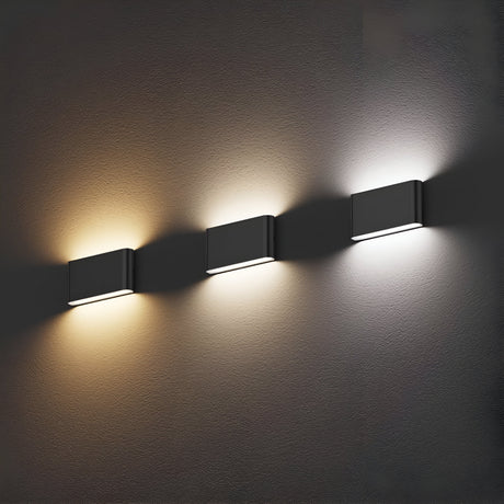 Three Jens-1 LED Outdoor Wall Lights in black are mounted on a textured surface. Each IP65-rated light emits a different color: warm yellow on the left, soft white in the middle, and cool white on the right, casting subtle shadows below them.