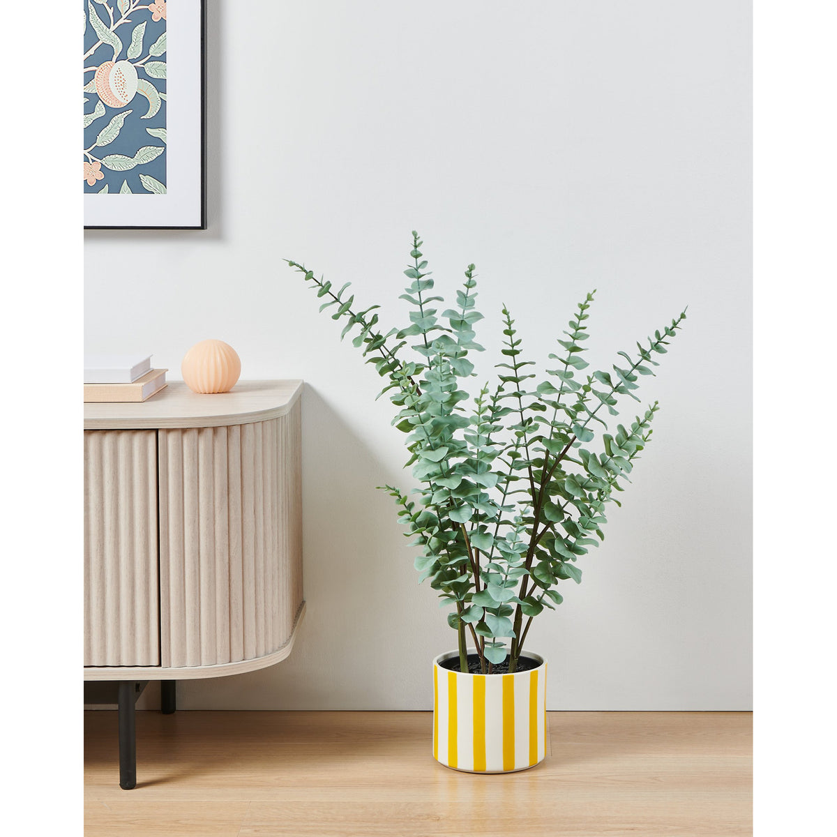 Jinjiti 14cm ceramic plant pot with white and yellow vertical stripes, holding a potted eucalyptus plant beside a wooden sideboard in a living room setting.