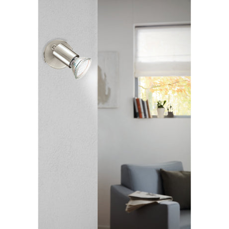 Josmanne Wall Spotlight in satin nickel with round backplate and pivoting cylindrical head, shown mounted on a white wall in a living room setting.