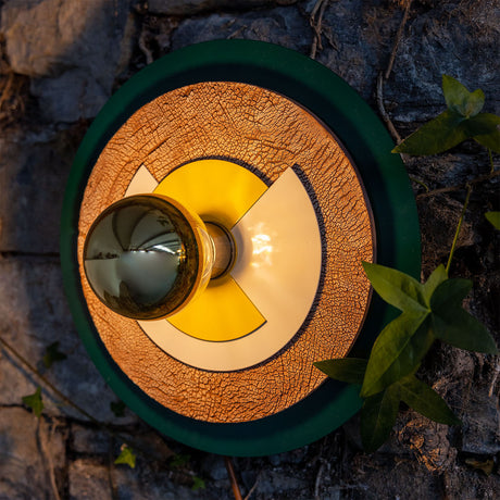 Jupiter Modern Wall Light with layered round discs in textured brass and cream, a dark green outer ring, and a brass bulb holder, mounted on a stone wall with leaves.
