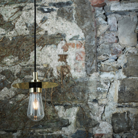Kai Glass Disc Pendant Light IP65 with a flat brass disc shade over a clear teardrop glass shade, exposed bulb, and black cable, hanging against a stone wall.
