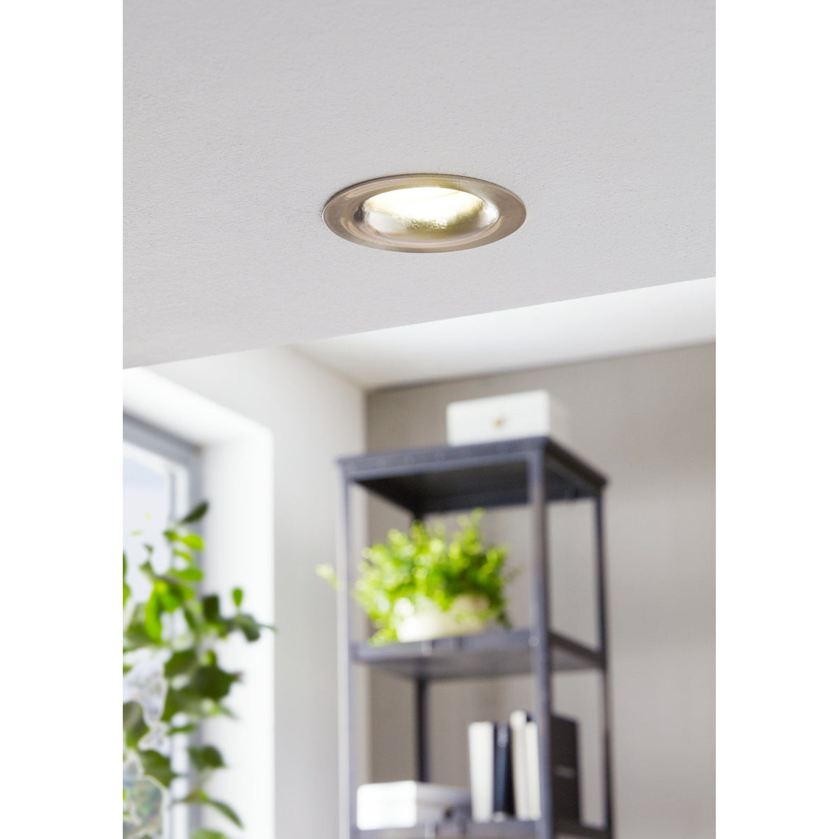 Kamarne LED Downlight recessed into a white ceiling, satin nickel trim with ribbed reflector, shown lit above a dark shelving unit with plants and books.