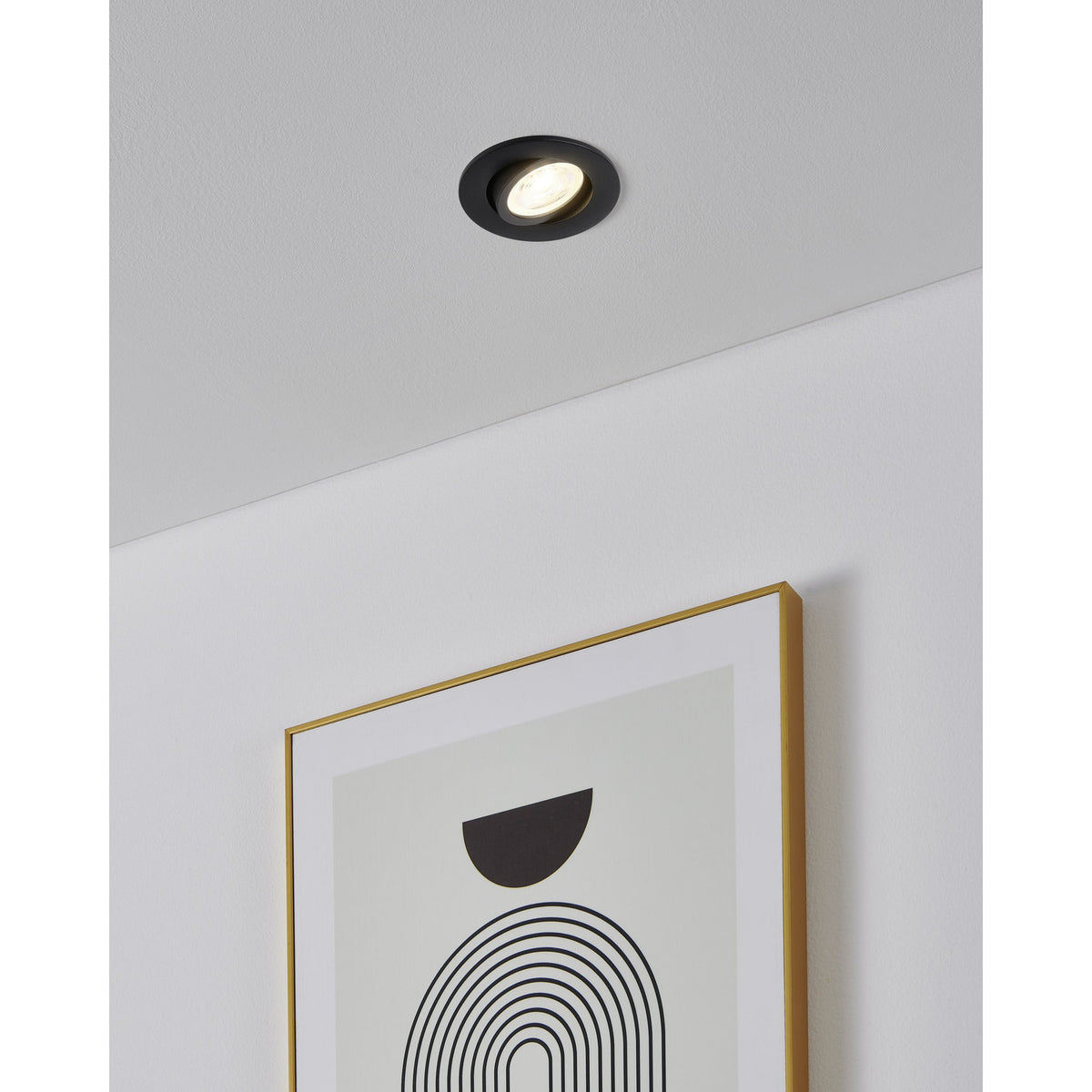 Round black aluminium recessed downlight from the Kamarne set installed in a ceiling, with tilting inner ring angled above a framed abstract print on a white wall.