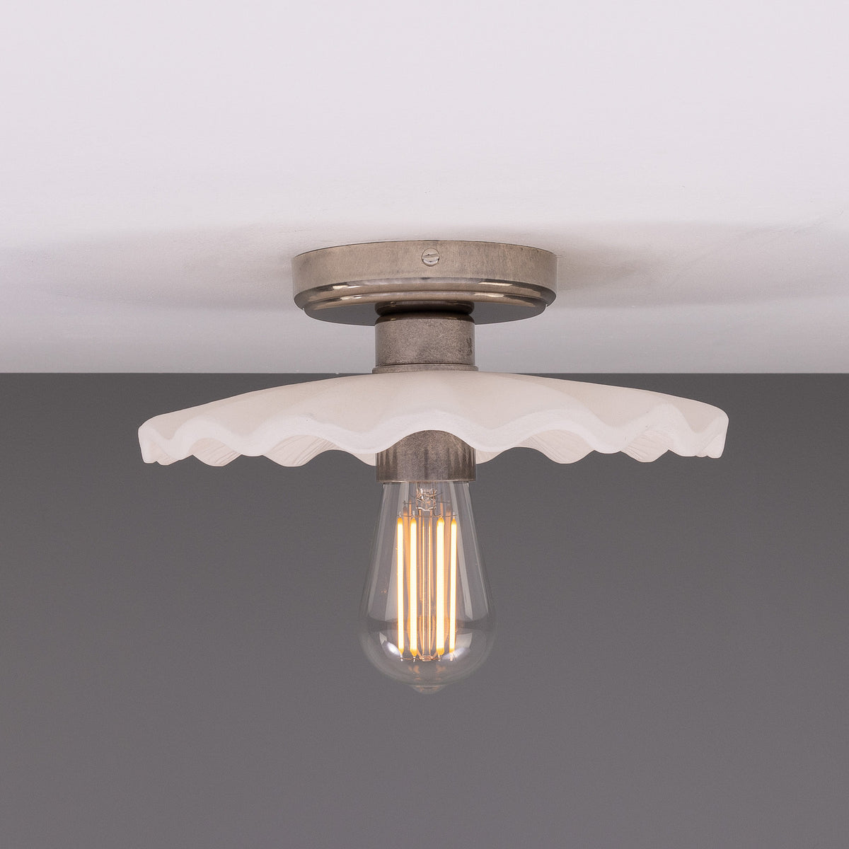 Kapok ceramic ceiling light with a scalloped, matt white striped shade and aged metal lampholder, shown lit with a vintage filament bulb against a grey background.