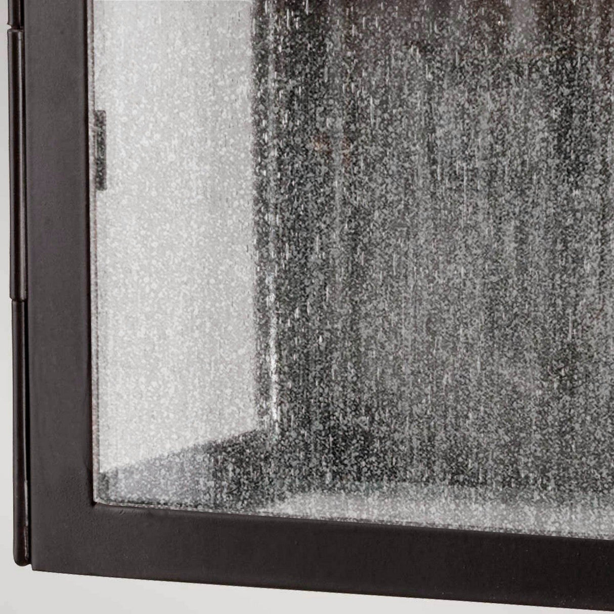 A close-up of the Kendal 2 Light Outdoor Wall Light in an old bronze finish reveals a textured glass panel within a black metal frame, reminiscent of classic lantern design. The frosted, speckled appearance of the glass partially obscures the view behind it, evoking an old bronze charm.