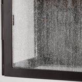 A close-up of the Kendal 2 Light Outdoor Wall Light in an old bronze finish reveals a textured glass panel within a black metal frame, reminiscent of classic lantern design. The frosted, speckled appearance of the glass partially obscures the view behind it, evoking an old bronze charm.