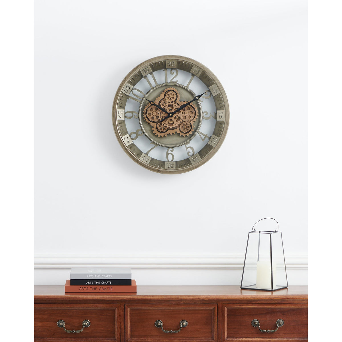 Kenema 46cm round wall clock with skeleton face, cut-out Arabic numerals, exposed antique gold gears and metal frame, shown above a wooden sideboard with candle lantern and books.