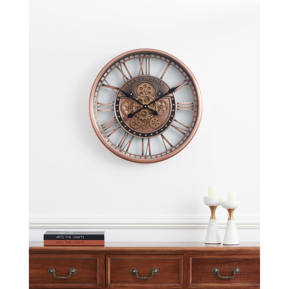Kenema Wall Clock 53.5cm in antique copper, round skeleton frame with Roman numerals and exposed gear centre, shown above a wooden sideboard with candlesticks and books.