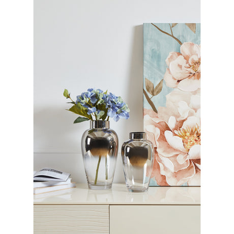 Kenyasi Glass Vase 27cm, a tall shouldered vase with mirrored silver finish fading to clear glass, shown with blue hydrangeas on a sideboard beside a smaller matching vase and floral artwork.