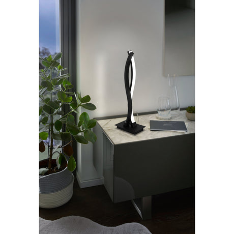Keselle LED table lamp in black with twisted steel arms on a square base, shown on a cabinet beside a mirror and potted plant.