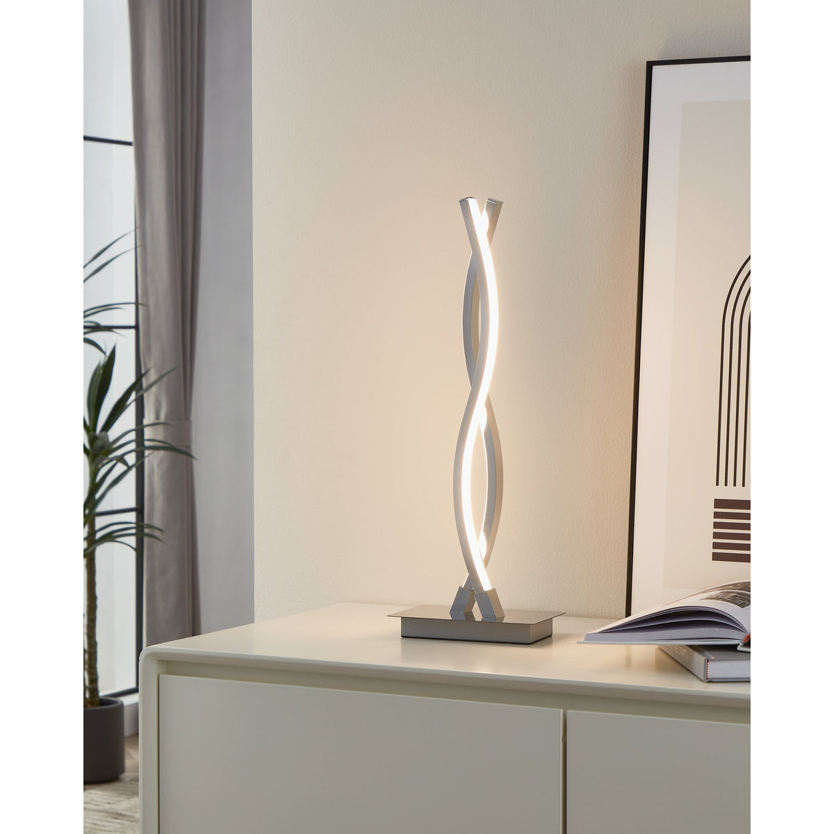 Keselle LED table lamp with intertwined wave-shaped chrome arms on a rectangular base, glowing warm white, shown on a sideboard in a living room setting.