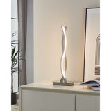 Keselle LED table lamp with intertwined wave-shaped chrome arms on a rectangular base, glowing warm white, shown on a sideboard in a living room setting.