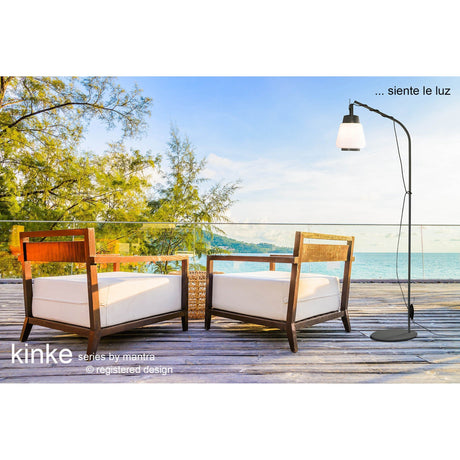 Two wooden lounge chairs with white cushions rest on a deck overlooking the ocean. Nearby stands the Kinke Floor Lamp With Hook - Anthracite, a sleek outdoor luminaire. Trees and blue sky fill the background. Text: ...siente la luz and kinke series by mantra.