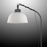 The Kinke Floor Lamp With Hook - Anthracite features a ribbed white dome shade suspended from a curved black metal stand, with its cord wrapped around the upper section, set against a grayscale gradient background.