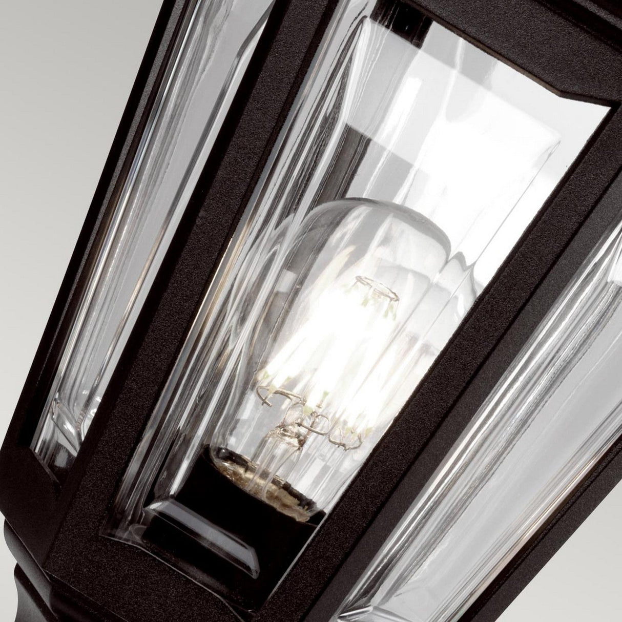 Close-up of the Kinsale 1 Light Pedestal/Porch Light in black, featuring a vintage-style design with a clear glass panel. Inside, an illuminated light bulb reveals its intricate filament design. This weather-resistant outdoor lighting piece stands elegantly against a plain, light gray background.