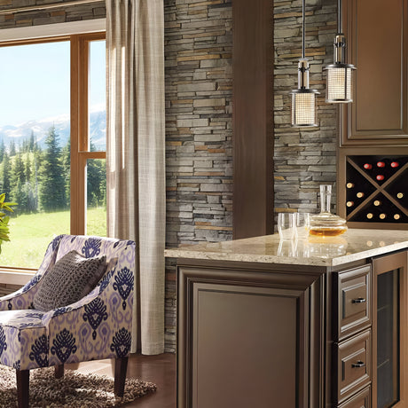 A cozy living room features a stone wall and a wine cabinet illuminated by an Ahrendale 1 Light Mini Pendant in grey. A patterned armchair sits beside a large window showcasing views of trees and mountains. Industrial-inspired decanters and glasses adorn the countertop, completing the inviting atmosphere.