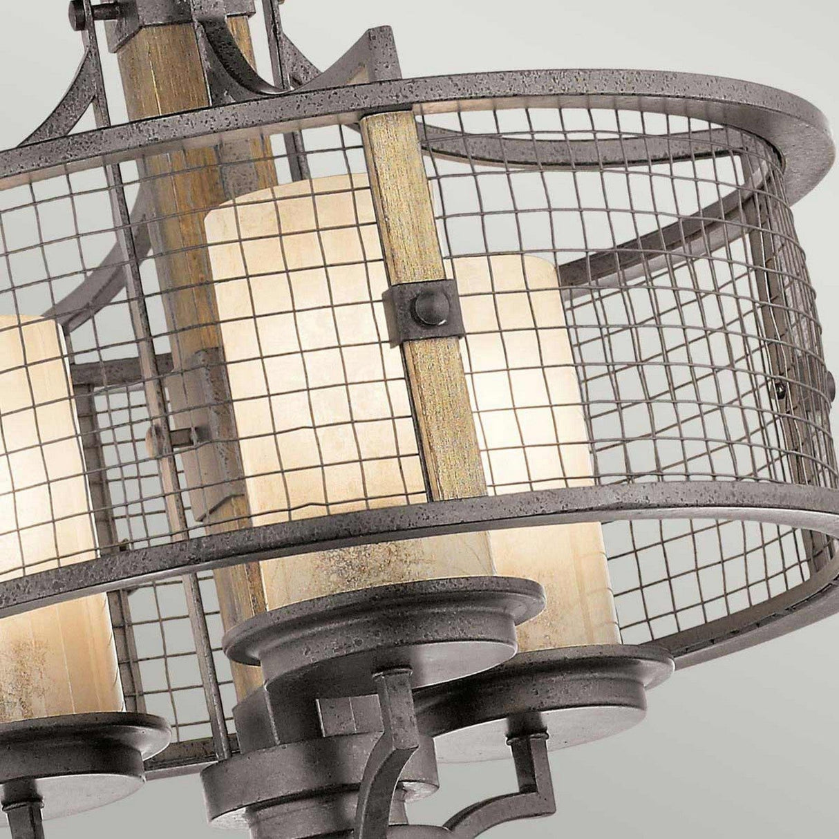 A close-up of the Ahrendale 3 Light Duo-Mount Chandelier in Grey highlights its rustic elegance with an industrial-style design. It features a cylindrical metal mesh cage that encloses three frosted glass, candle-shaped bulbs, while the frame showcases a vintage finish, all illuminated.