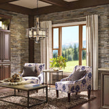 A cozy living room with stone brick walls exudes rustic elegance, featuring two patterned armchairs, a wooden coffee table, and a large window framing a view of the green landscape and mountains. The Ahrendale 3 Light Duo-Mount Chandelier in Grey introduces a modern flair from above, while a plant enhances the inviting atmosphere.