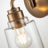 A close-up shot of the Aivian 1 Light Wall Light in weathered brass reveals a clear glass shade with a glowing filament bulb inside. The brushed metal finish imparts an industrial feel, enhancing its warm, modern aesthetic against a plain background.