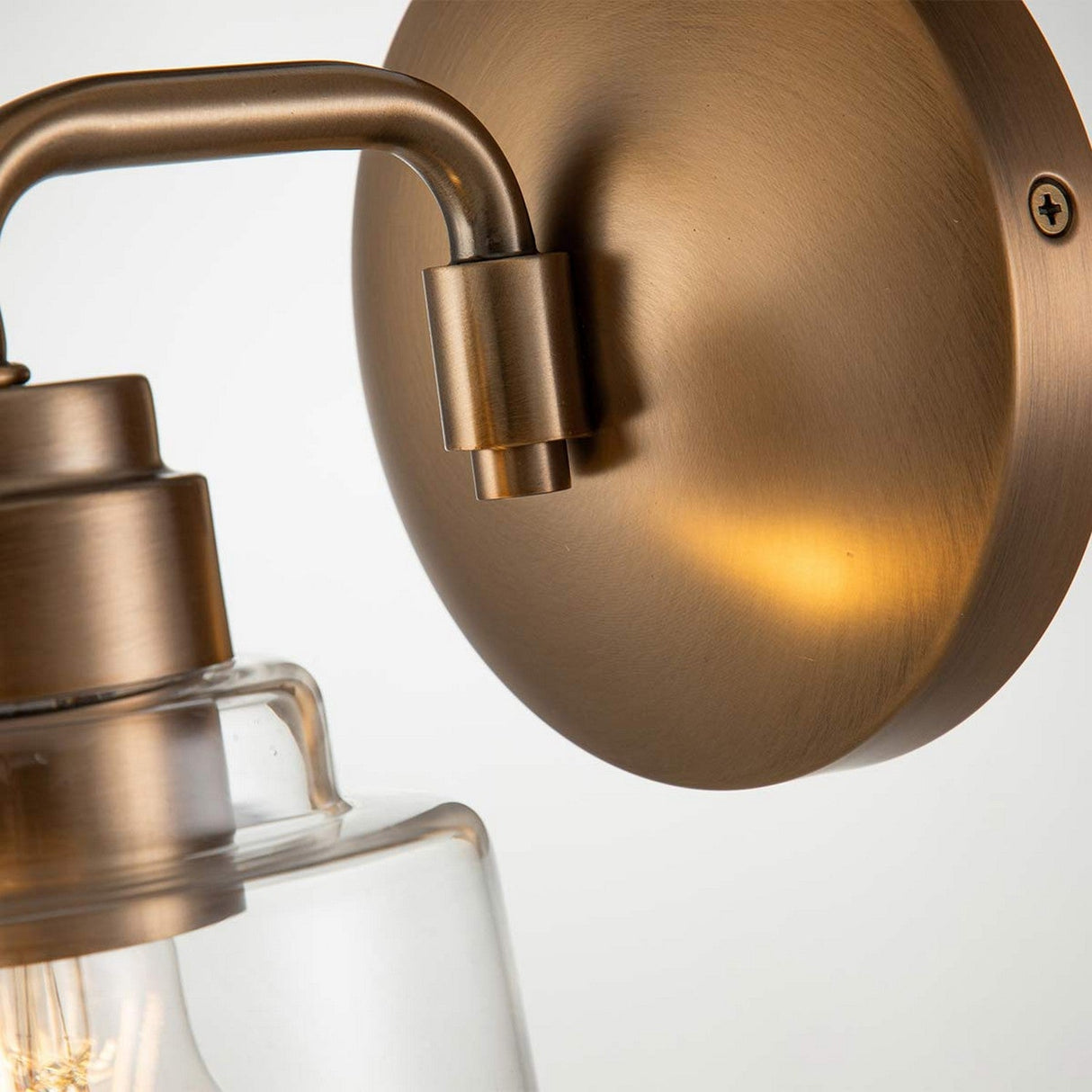 Close-up of the Aivian 1 Light Wall Light - Weathered Brass, featuring a wall-mounted fixture with a clear glass shade. Its smooth, brushed finish lends an industrial edge, allowing partial visibility of the bulb within its weathered brass design.