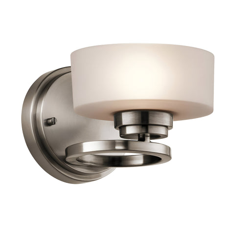 The Aleeka 1 Light Wall Light - Silver features a modern design with a brushed nickel finish and a round frosted glass shade. Its circular base and minimalist metallic accents enhance contemporary interiors, making it a stylish choice for indoor lighting.