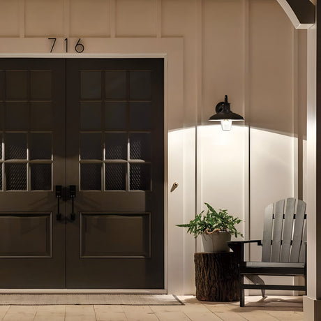 A well-lit entrance features dark double doors, each with nine glass panes. An Allenbury Outdoor 1 Light Medium Wall Lantern in black (IP44 rated) casts light on a potted plant resting on a wooden stump beside a black Adirondack chair. Above the doors, the number 716 is displayed, showcasing weather-resistant lighting elegance.