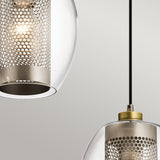 The Asher 3 Light Pendant Rectangle - Silver creates a sleek, modern ambiance with its three contemporary pendant lights. It features cylindrical mesh metal shades encased in clear glass exteriors, each suspended from a black cord. These lights cast a warm, soft glow against the plain, light gray surface for an industrial lighting vibe.