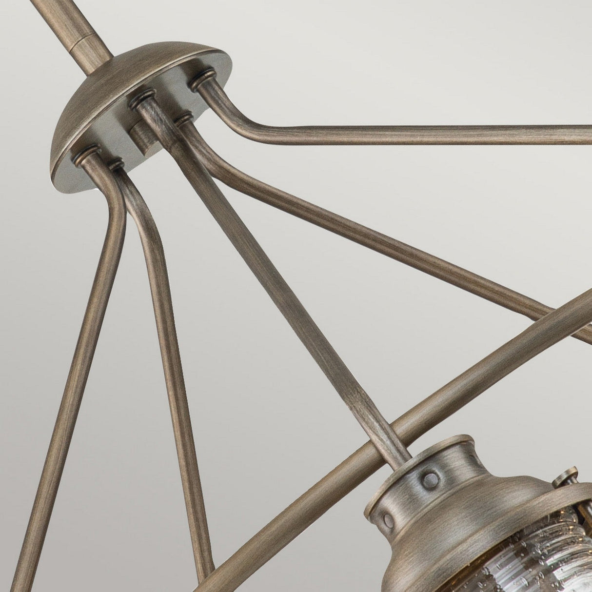 Close-up of the Ashland Bay Outdoor 5 Light Chandelier, showcasing its metallic industrial style with a burnished bronze finish, intricate geometric rod designs, and a textured seeded glass light fixture. The background is a plain light gray.