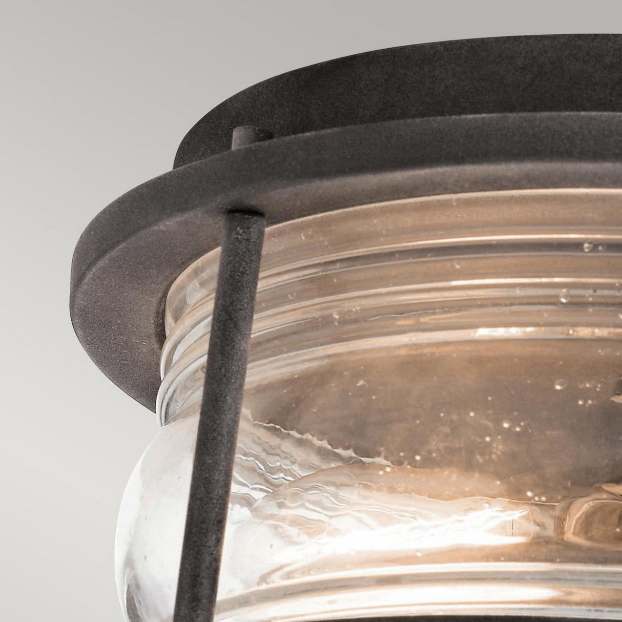 Close-up of the Ashland Bay Outdoor 2 Light Outdoor Ceiling Flush in weathered zinc, showcasing a black metal and glass lantern with a circular design. The ribbed texture of the clear seeded glass encases the light bulb, exuding an elegant ambiance. This modern fixture is set against a plain gray background.
