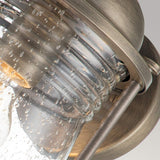 Close-up of the Ashland Bay Outdoor 1 Light Medium Wall Lantern showcasing its vintage-style design with a burnished bronze metal frame and clear seeded glass cover featuring small air bubbles. The fixture's weathered, antique finish contributes to its rustic appearance against a neutral background.