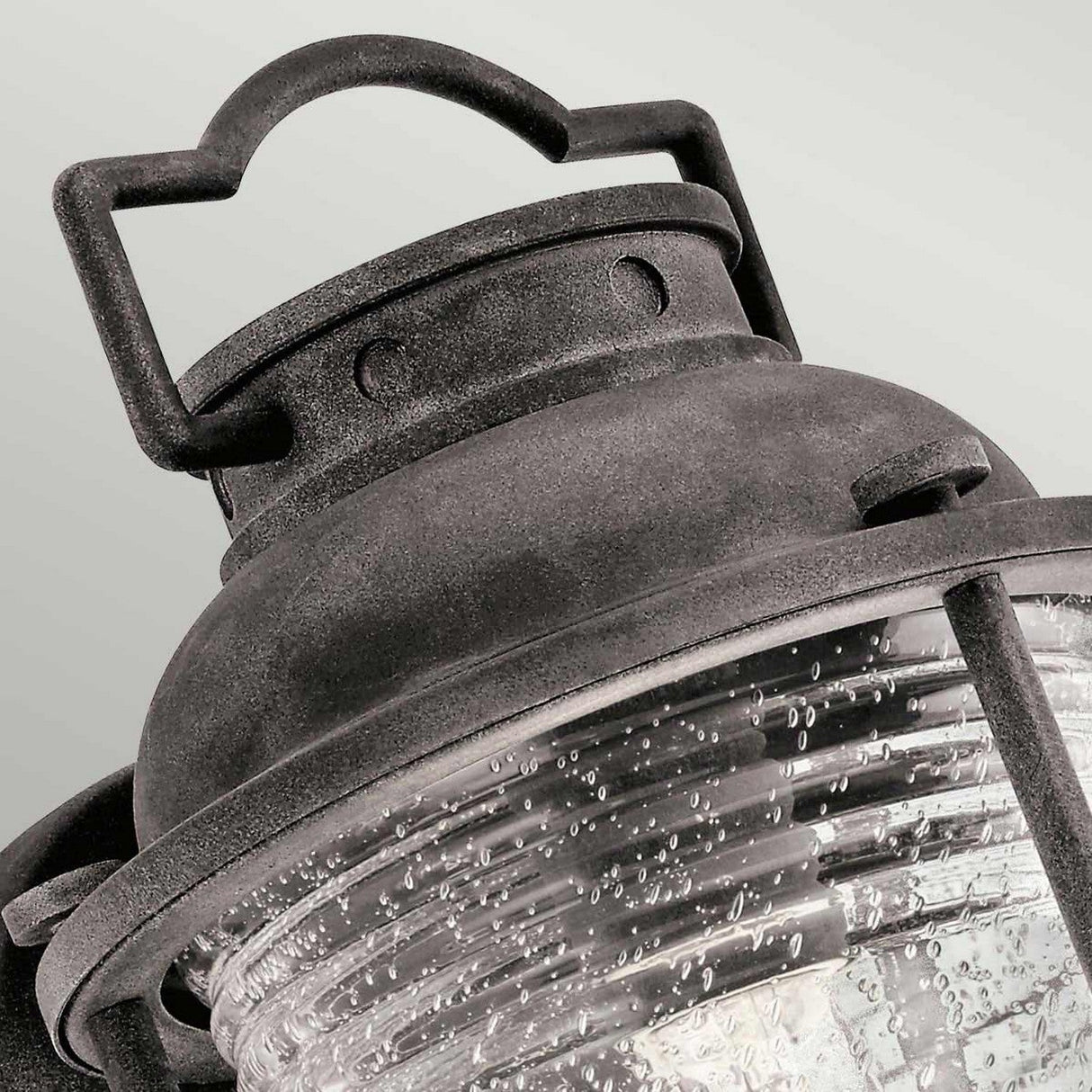 Close-up of the Ashland Bay Outdoor 1 Light Medium Wall Lantern, designed in a vintage style with a weathered zinc finish and seeded glass. It features a curved glass casing with small bubbles that enhance its rustic appearance. The lantern includes a handle at the top for easy carrying or hanging.