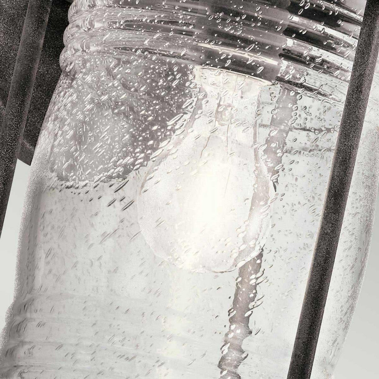 The detailed view of the Ashland Bay Outdoor 1 Light Medium Wall Lantern in Weathered Zinc showcases its stunning seeded glass, adorned with gentle textures and bubbles that accentuate the luminous bulb within. The weathered zinc frame provides a rustic contrast to the sleek, reflective glass surface.