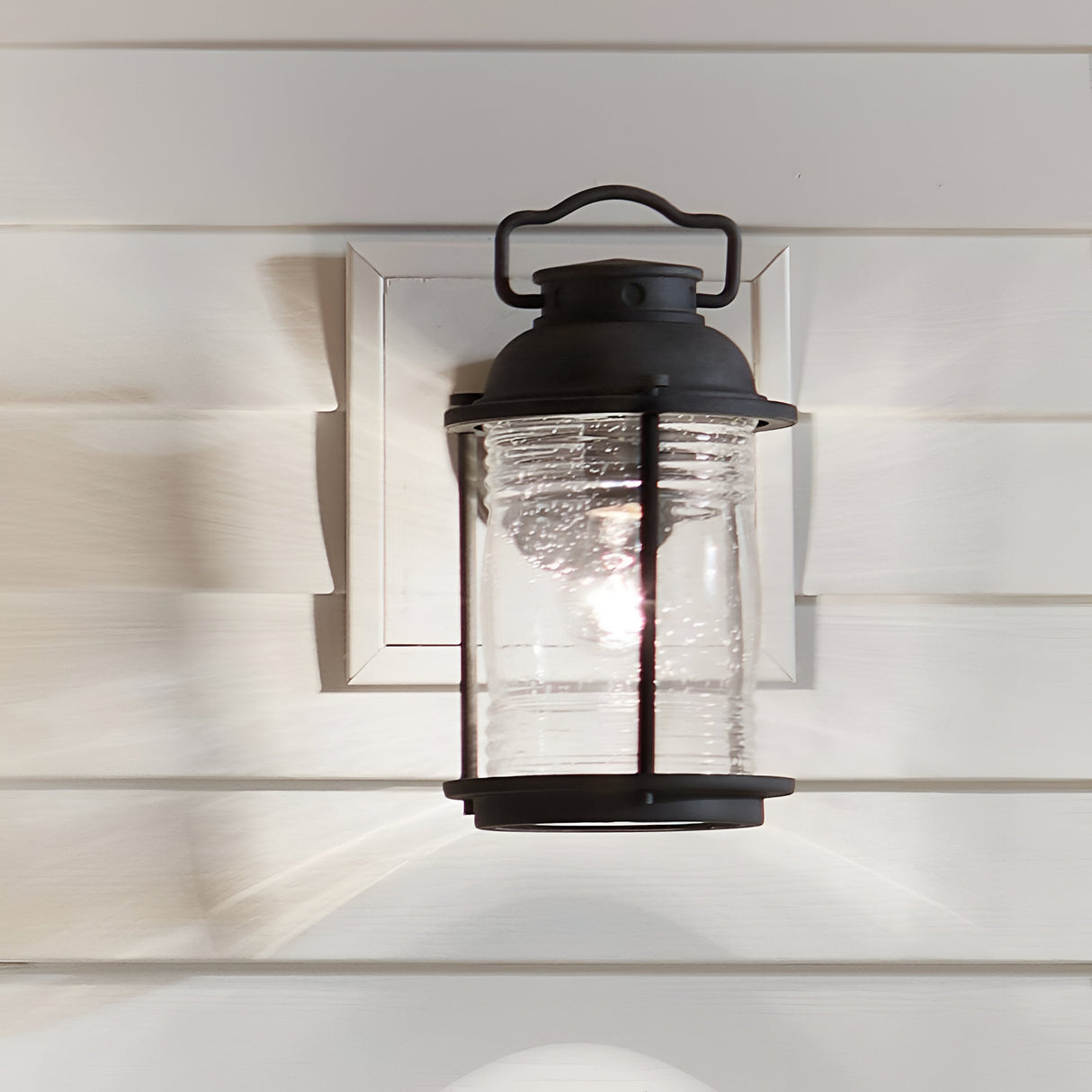 The Ashland Bay Outdoor 1 Light Medium Wall Lantern in a weathered zinc finish and featuring clear seeded glass panels is mounted on a light-colored siding wall. The illuminated lantern casts a soft glow, creating intricate light patterns on the wall.