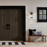 A modern exterior showcases dark double doors outlined in black against a white paneled wall. To the right, beneath a window framed with weathered zinc, is a wooden bench adorned with black and gray cushions. A striped rug is placed in front of the doors, while an Ashland Bay Outdoor 1 Light Medium Wall Lantern in Weathered Zinc hangs above, featuring seeded glass.