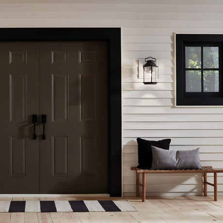 A modern exterior showcases dark double doors outlined in black against a white paneled wall. To the right, beneath a window framed with weathered zinc, is a wooden bench adorned with black and gray cushions. A striped rug is placed in front of the doors, while an Ashland Bay Outdoor 1 Light Medium Wall Lantern in Weathered Zinc hangs above, featuring seeded glass.