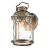 The Ashland Bay Outdoor 1 Light Small Wall Lantern in burnished bronze exudes vintage charm with its clear, ribbed glass shade and LED Edison bulb that emits a warm glow. It features a top handle and is mounted on a circular base for added appeal.