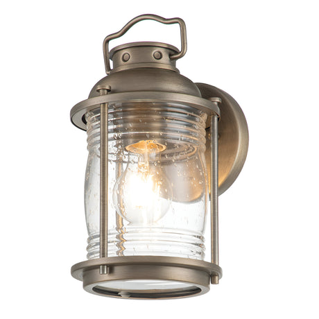 The Ashland Bay Outdoor 1 Light Small Wall Lantern in burnished bronze exudes vintage charm with its clear, ribbed glass shade and LED Edison bulb that emits a warm glow. It features a top handle and is mounted on a circular base for added appeal.