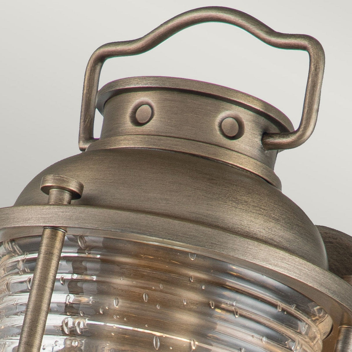 Close-up of the Ashland Bay Outdoor 1 Light Small Wall Lantern in burnished bronze. The top section showcases a handle and rivet details, while the lower part features ribbed glass housing LED Edison bulbs, creating a warm and inviting glow.