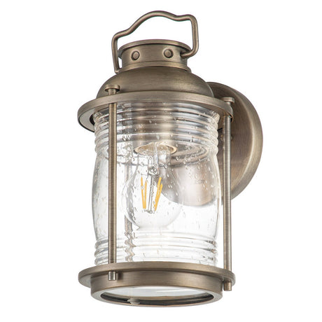 Introducing the Ashland Bay Outdoor 1 Light Small Wall Lantern in Burnished Bronze, which elegantly combines vintage charm with modern design. This wall lantern showcases a clear glass encasement that highlights a visible LED Edison bulb, all mounted thoughtfully on a round base with an appealing handle-like accent on top.