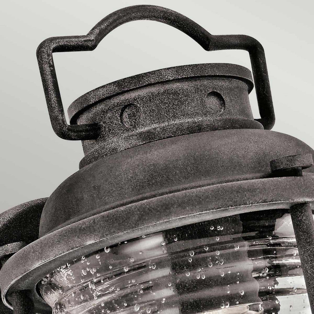 Close-up of the Ashland Bay Outdoor 1 Light Small Wall Lantern in weathered zinc, featuring a clear, ridged glass cover. Its decorative handle highlights a charming Colonial design, giving the lantern a rustic appearance.