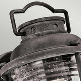 Close-up of the Ashland Bay Outdoor 1 Light Small Wall Lantern in weathered zinc, featuring a clear, ridged glass cover. Its decorative handle highlights a charming Colonial design, giving the lantern a rustic appearance.