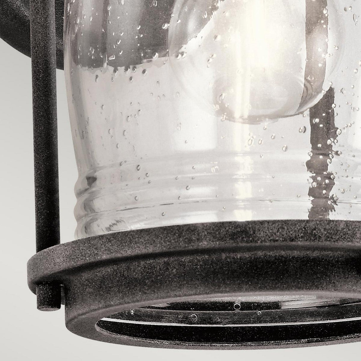 Close-up of the Ashland Bay Outdoor 1 Light Small Wall Lantern showcasing a colonial design. This lantern features a clear, seeded glass enclosure with charming bubbles that beautifully complements the weathered zinc metal frame encasing the lamp. A light bulb is visible inside.